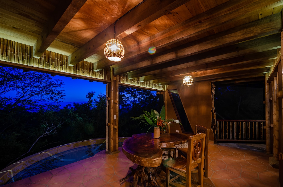 An indoor dining area features a large wooden table surrounded by mismatched chairs, softly illuminated by hanging woven light fixtures. The room opens to a lush forest view, enhancing the connection to nature with earthy tones and natural materials throughout.