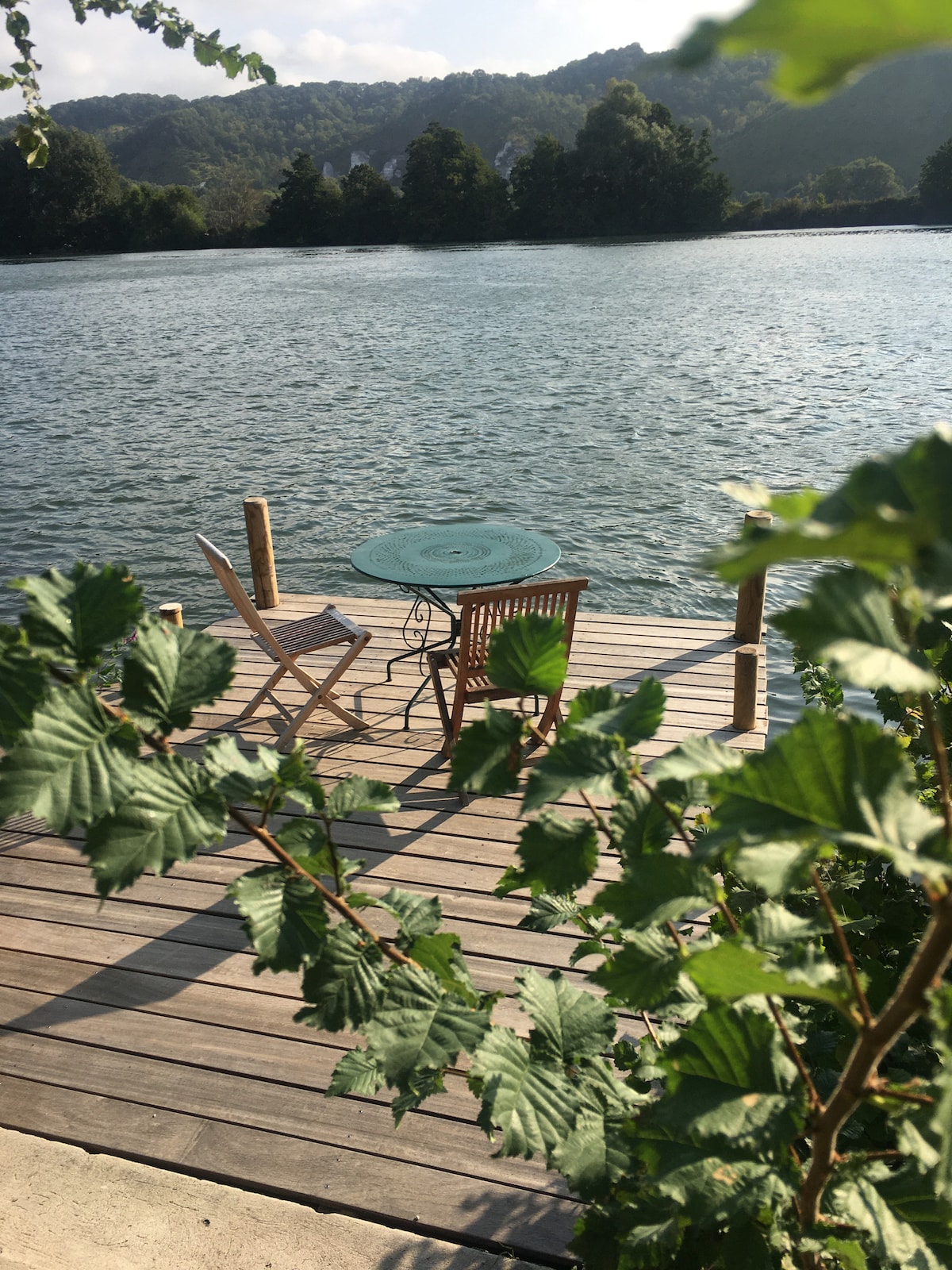 A wooden dock extends over the water, featuring a small circular table surrounded by two chairs. Lush greenery in the foreground frames the scene, while calm water and distant green hills provide a serene backdrop.