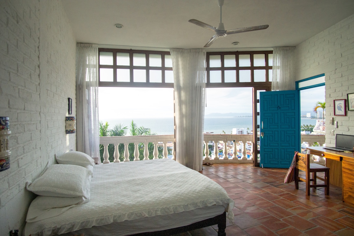 A spacious bedroom features a large bed with white linens. Sunlight filters through multiple windows, offering views of the ocean. Fresh breezes can be felt throughout the room. A wooden desk and chair are positioned near the window, creating a bright workspace.