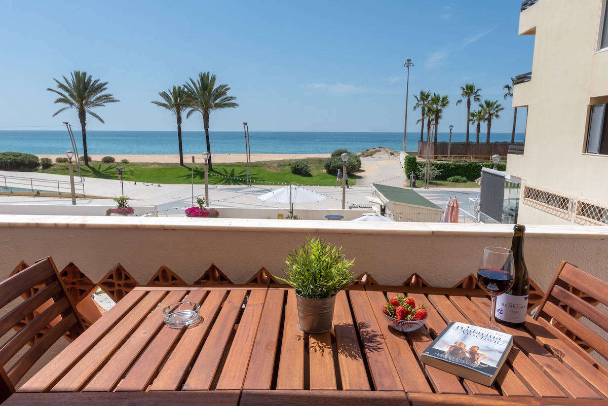 A wooden dining table is set on the balcony, overlooking the beachfront of Quarteira. Fresh strawberries and a glass of red wine are placed on the table, alongside a book. Palms frame the view of the sandy beach and calm sea under a clear blue sky.