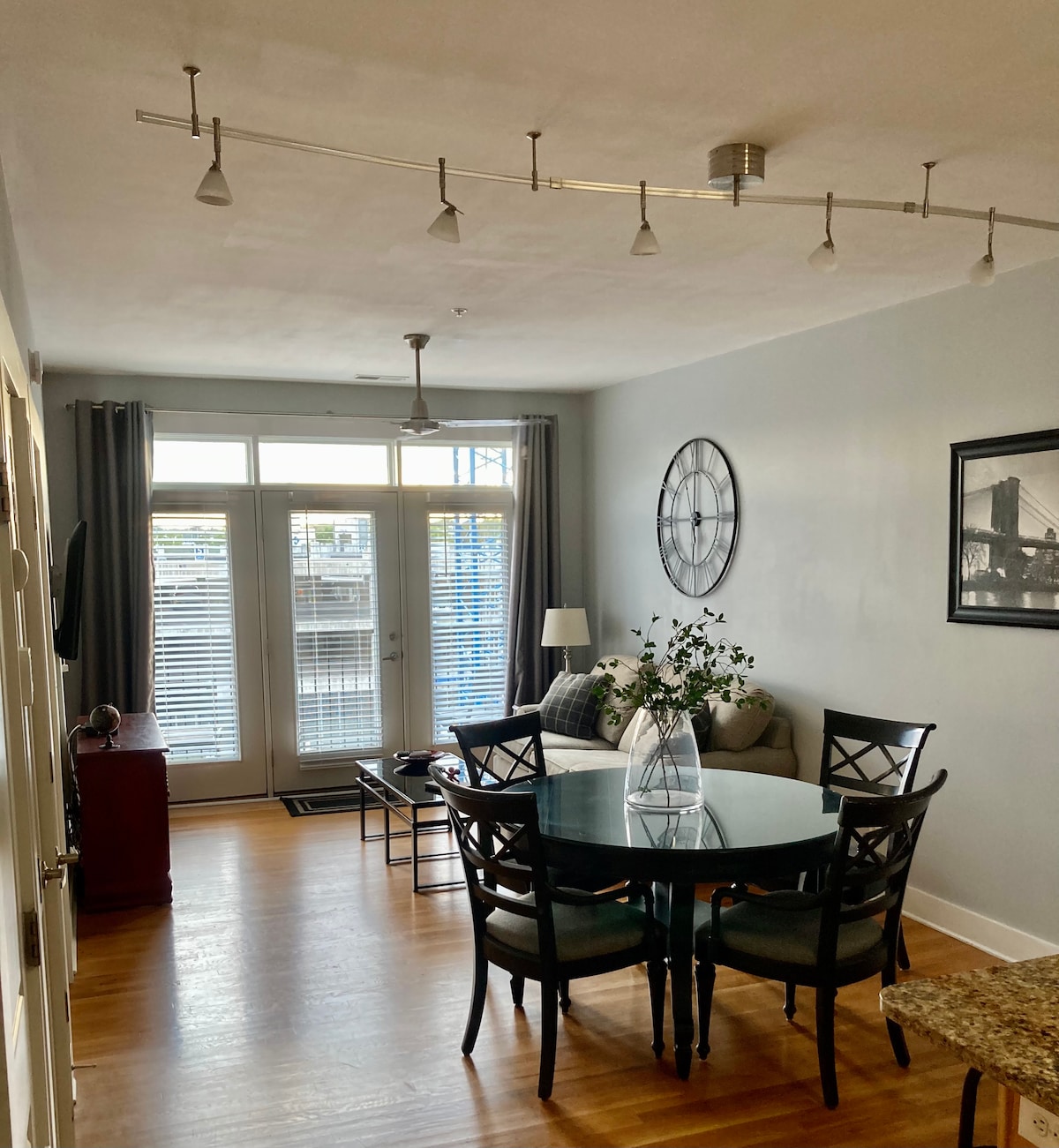 A dining area is highlighted by a glass table surrounded by four chairs. A light fixture runs along the ceiling, providing illumination. Large windows with light curtains allow natural light to fill the space, which features a comfortable sofa and a decorative wall clock.