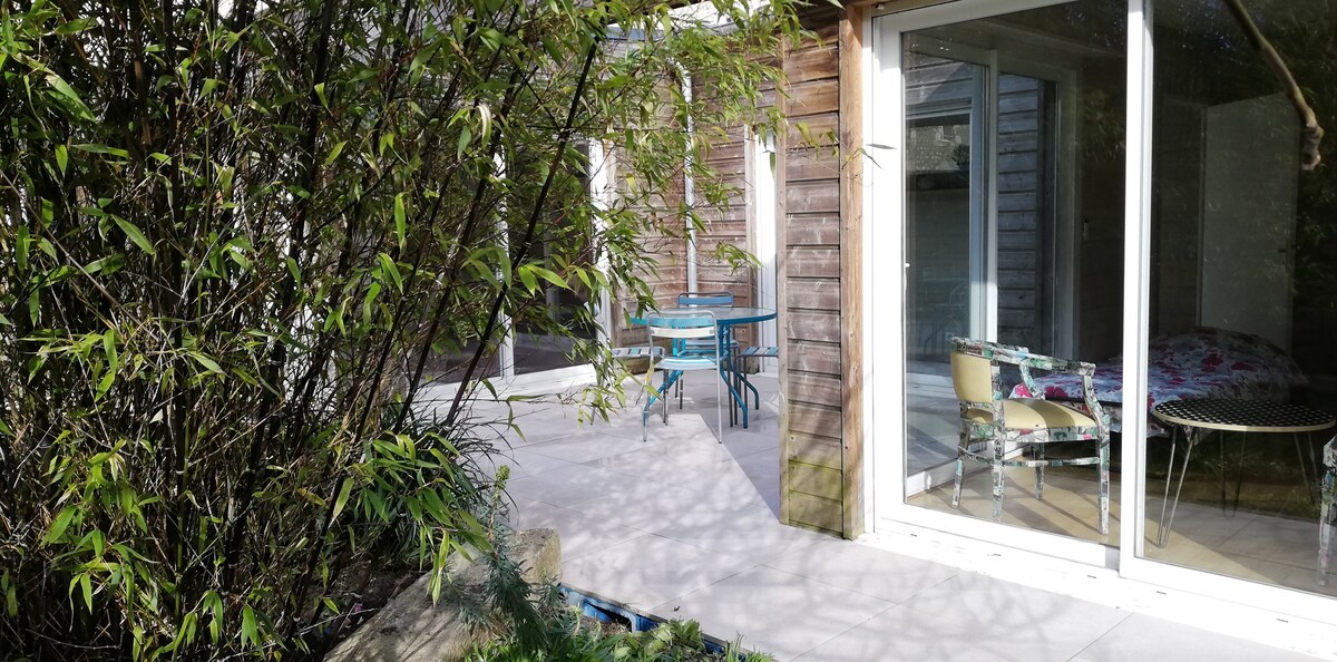 An external view of the home, featuring a serene outdoor seating area adorned with a round table and colorful chairs. Lush green bamboo plants offer a natural backdrop, and large glass doors provide access to the interior, allowing for plenty of natural light.