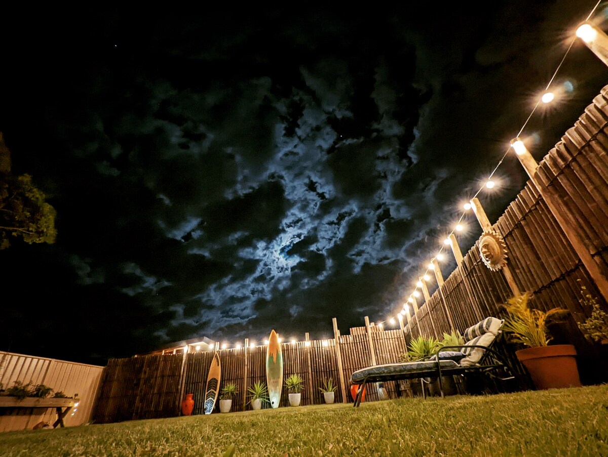The spacious backyard is illuminated by string lights against a night sky filled with clouds. Two colorful surfboards are positioned near the wooden fence, and various potted plants are arranged along the perimeter. A lounge chair sits in the foreground, inviting relaxation.