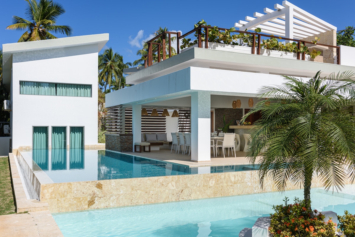 A modern villa is showcased with sleek architectural features and clean lines. An inviting swimming pool reflects the surrounding palm trees, while an open terrace with dining options overlooks the water. The exterior is accented by beautiful greenery and natural stone elements.