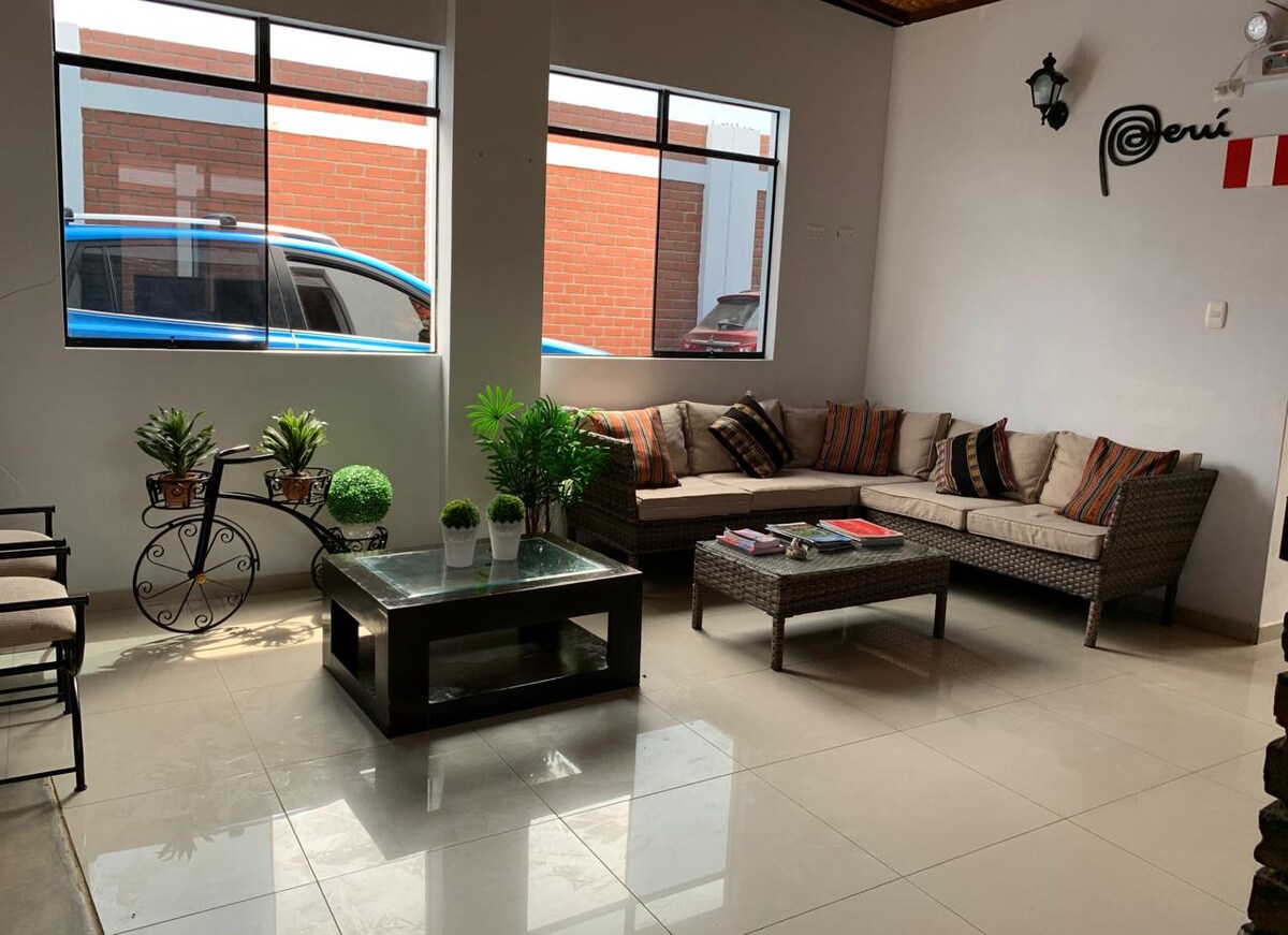 A welcoming lounge area features a comfortable sectional sofa adorned with decorative cushions. A glass coffee table sits at the center, accompanied by small potted plants. Large windows provide natural light, creating a bright atmosphere in the space.