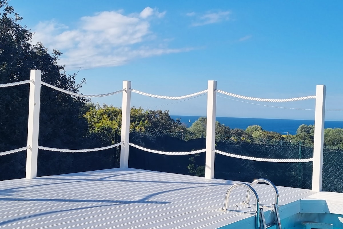 A spacious terrace with a protective railing offers expansive views of the azure sea and distant boats. Lush greenery surrounds the area, enhancing the outdoor setting. The terrace is adorned with smooth decking, providing a comfortable space for relaxation and enjoying the scenic landscape.