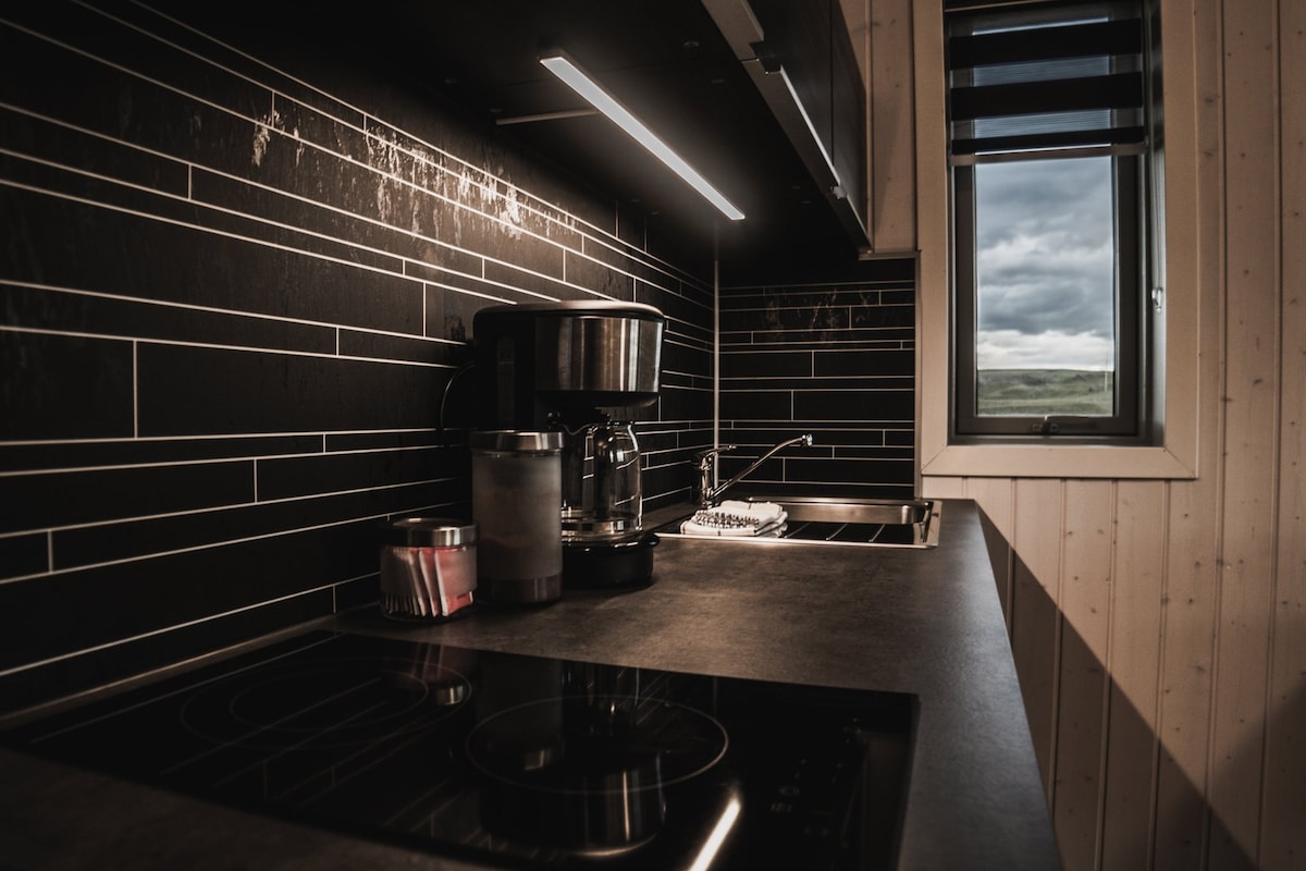 The kitchen displays a modern design with black tiled walls and sleek countertops. A coffee machine is positioned next to a small fridge, while a ceramic cooktop and sink are visible. Natural light enters through a window, highlighting the space.