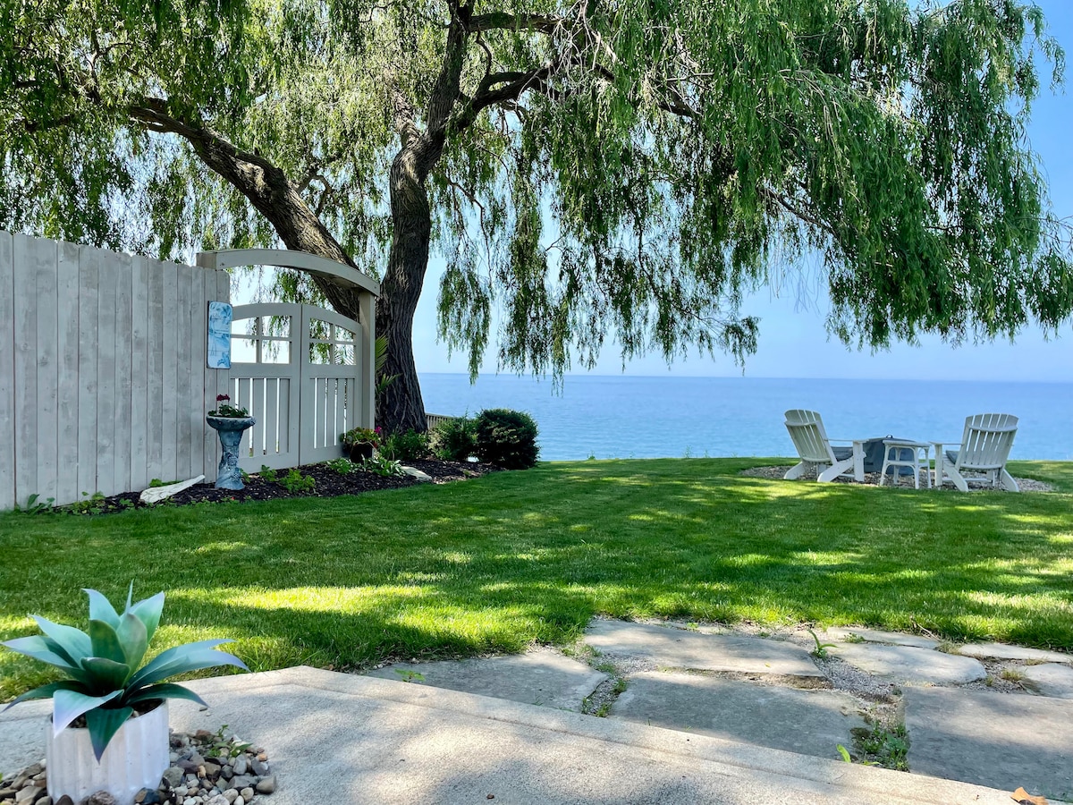 A spacious backyard is highlighted by a large, weeping willow tree providing shade over a grass area. Outdoor seating with lounge chairs is positioned facing Lake Erie, offering a view of the water. A peaceful atmosphere is accentuated by a small decorative planter in the foreground.
