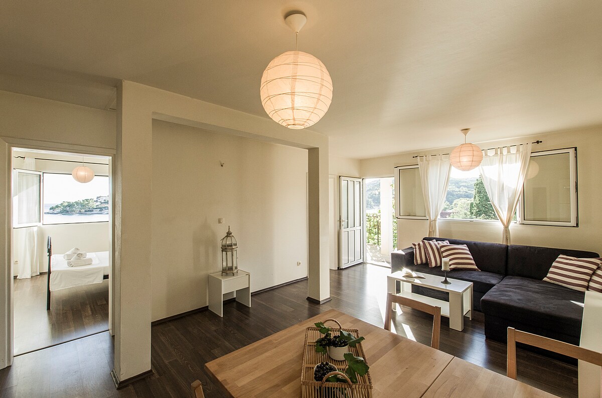 An open living area features a dark sectional sofa with striped cushions, complemented by a wooden dining table. Large windows allow natural light to fill the space, revealing views of the surrounding landscape. Soft pendant lighting hangs from the ceiling, enhancing the room's calm atmosphere.