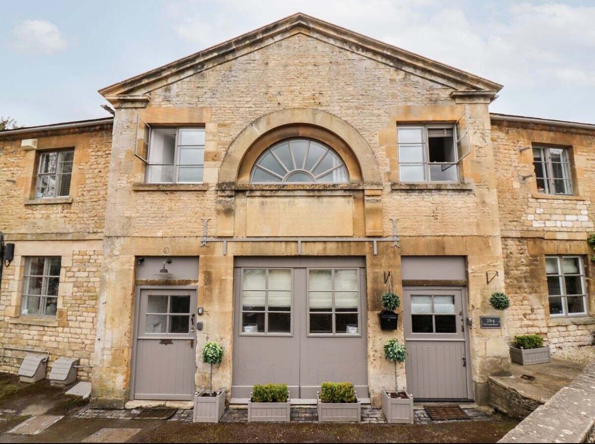 The front faƧade of The Coach House is showcased, featuring Cotswold stone construction with large windows. Two stylish doors welcome guests at the entrance, framed by potted plants. The structure exhibits traditional architectural elements alongside modern design features, contributing to its inviting appearance.