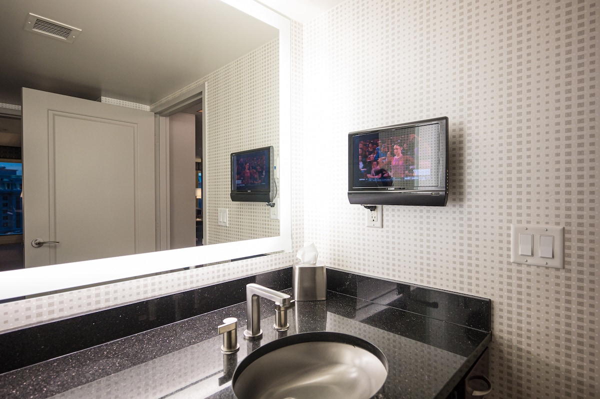 A modern bathroom features sleek black countertops paired with a minimalistic sink and faucet. A small flat-screen TV is mounted on the wall, with a large mirror reflecting the space. Soft lighting illuminates the area, enhancing its contemporary design.