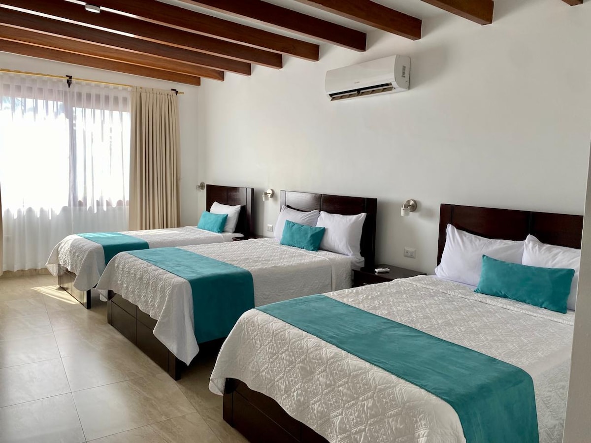 A spacious room features three beds with textured white linens and turquoise accents. Natural light floods the area through large windows, and air conditioning units are mounted above each bed. Soft, neutral tones create a calming environment, complemented by warm wooden beams overhead.