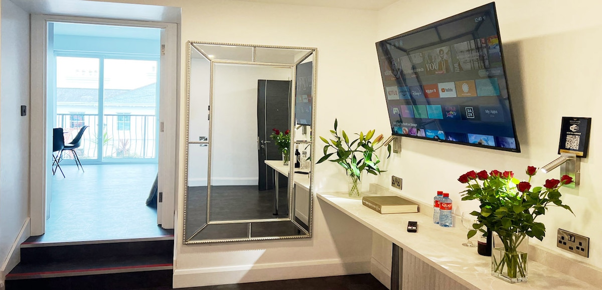 A sleek and modern interior is showcased, featuring a large mirror and a flat-screen television mounted on the wall. A decorative table is adorned with fresh flowers and bottled water. Light enters from a nearby balcony door, illuminating the open space.