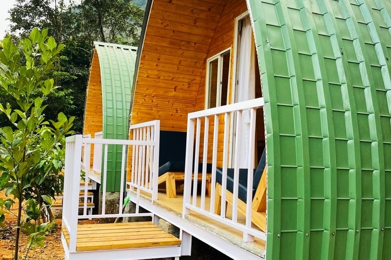 Two uniquely shaped cabins feature curved green exteriors and wooden accents. Each cabin has a private deck with seating options, inviting relaxation in a natural setting. Surrounding greenery enhances the tranquil atmosphere, while large windows provide ample natural light.