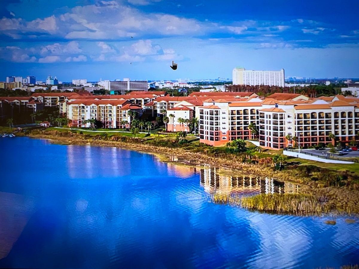 A panoramic view showcases a well-designed resort complex bordering a serene lake. The buildings reflect modern architecture with red-tiled roofs, surrounded by lush landscaping. The calm water mirrors the sky, enhancing the tranquil ambiance of the setting.