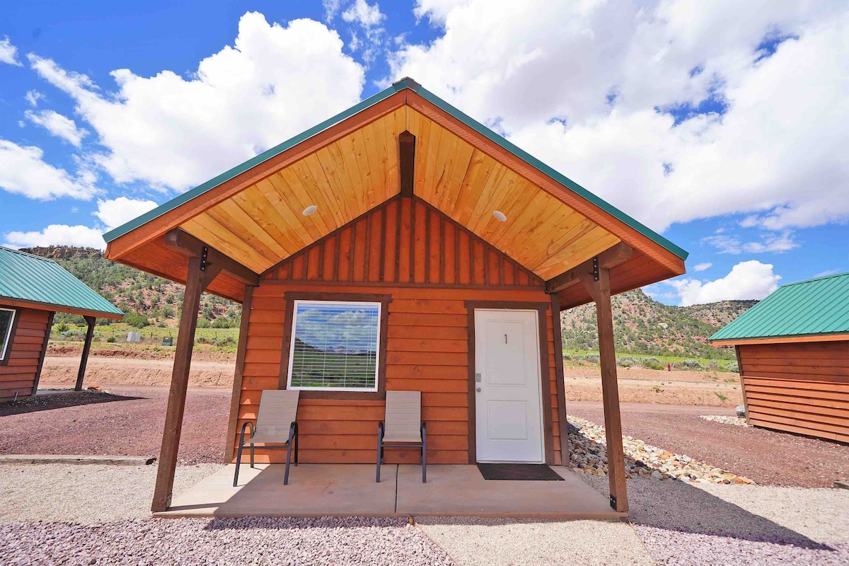 A rustic cabin exterior is shown with natural wood siding and a green metal roof. Two chairs are positioned on the porch, welcoming guests. The landscape features distant mountains and a partly cloudy sky, creating a serene atmosphere.