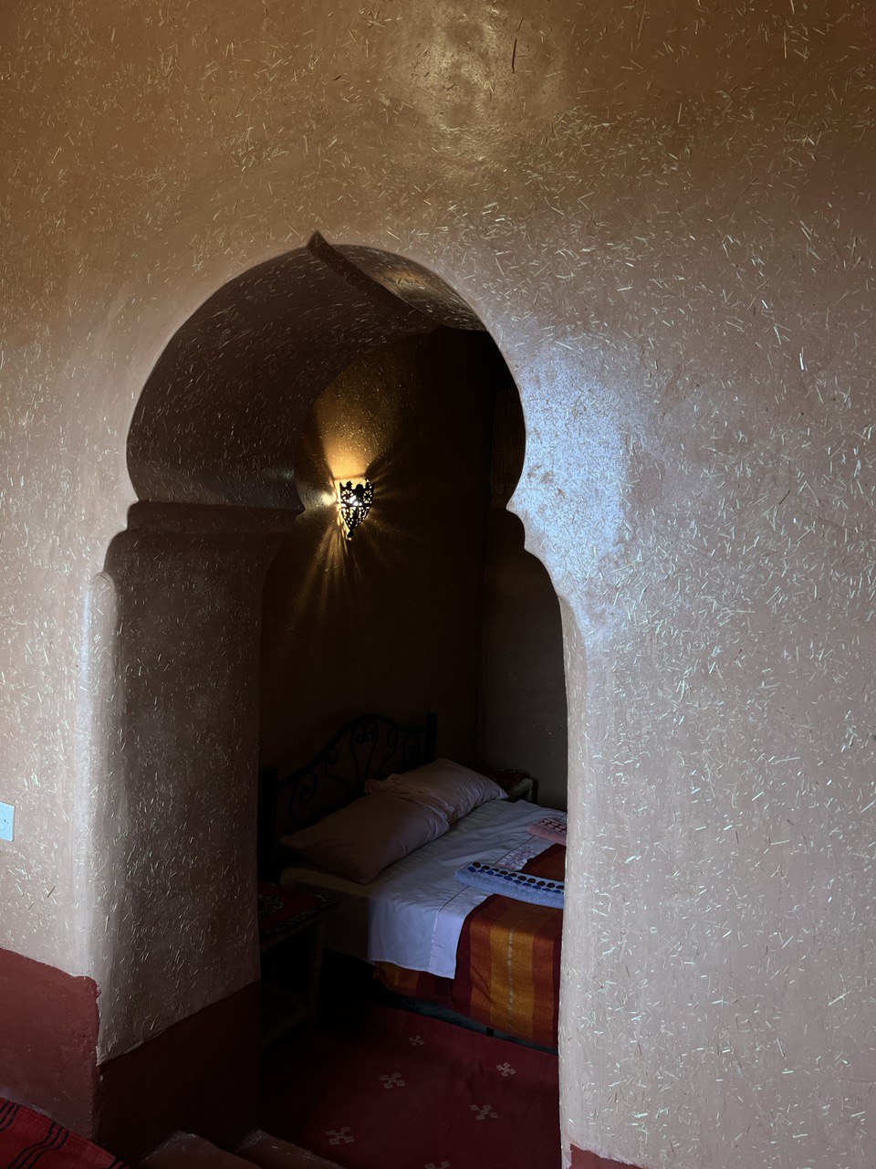 An interior view showcases an arched entryway leading to a cozy sleeping area. The earthy-toned walls, constructed with a textured finish, complement the soft lighting provided by a decorative wall sconce. A spacious bed with simple bedding is visible within the nook.