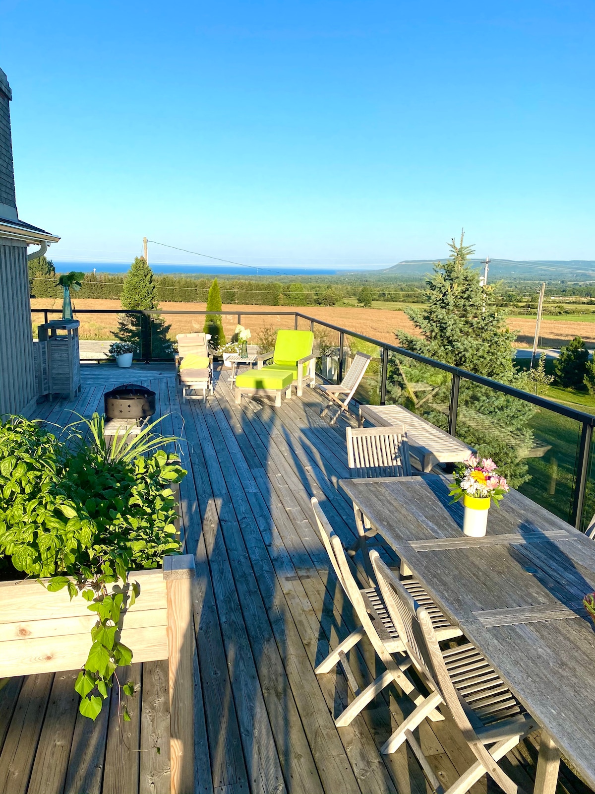 An upper deck with a wooden surface and glass railing overlooks a scenic landscape featuring greenery and distant blue waters. A dining table and chairs, along with a bright green lounge chair, are arranged for outdoor enjoyment. Potted plants add a touch of nature.