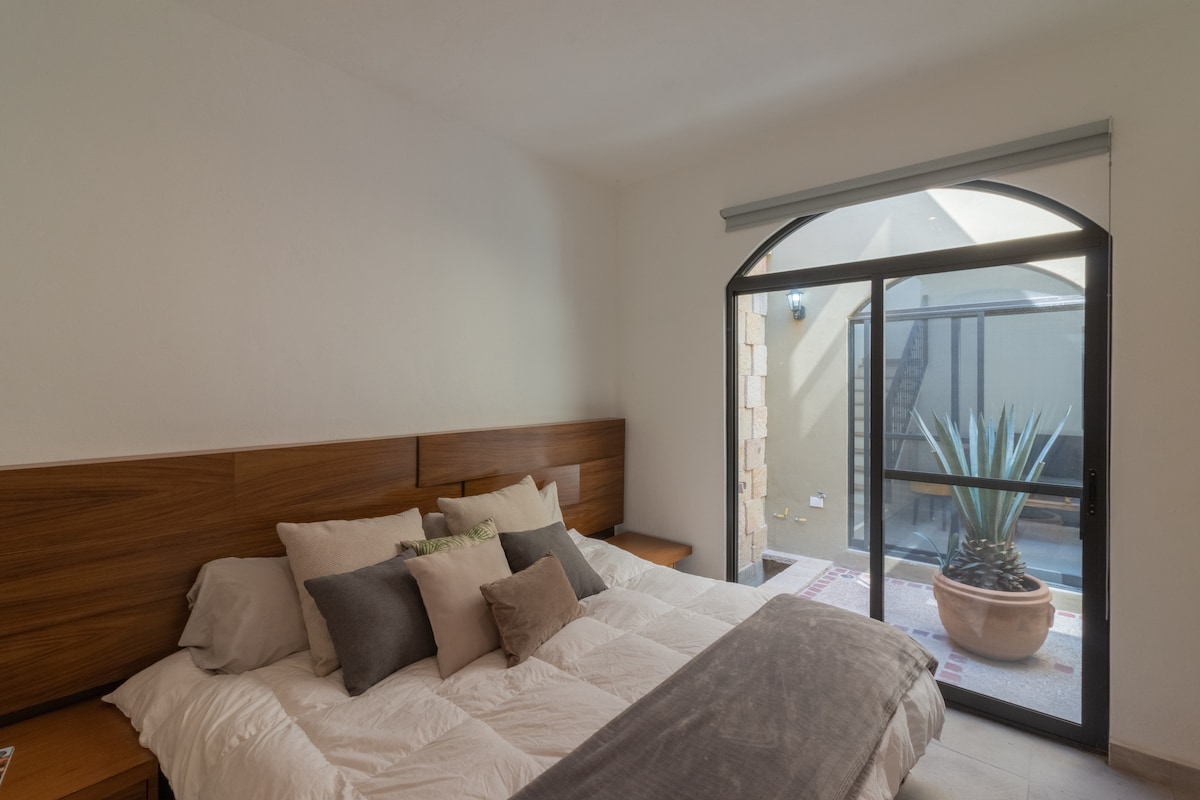 A spacious bedroom features a large bed with a light-colored quilt and decorative pillows. Natural light enters through a large arched window, which frames a view of a private courtyard with a potted plant. Simple wooden nightstands are positioned on either side of the bed.