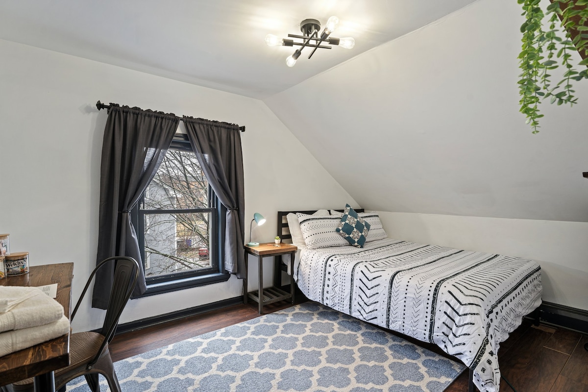 A well-lit bedroom features a spacious bed with a patterned duvet and coordinating decorative pillow. A handmade desk and nightstand are present, accompanied by a cozy rug beneath. Dark curtains frame the window, and a modern light fixture illuminates the room.