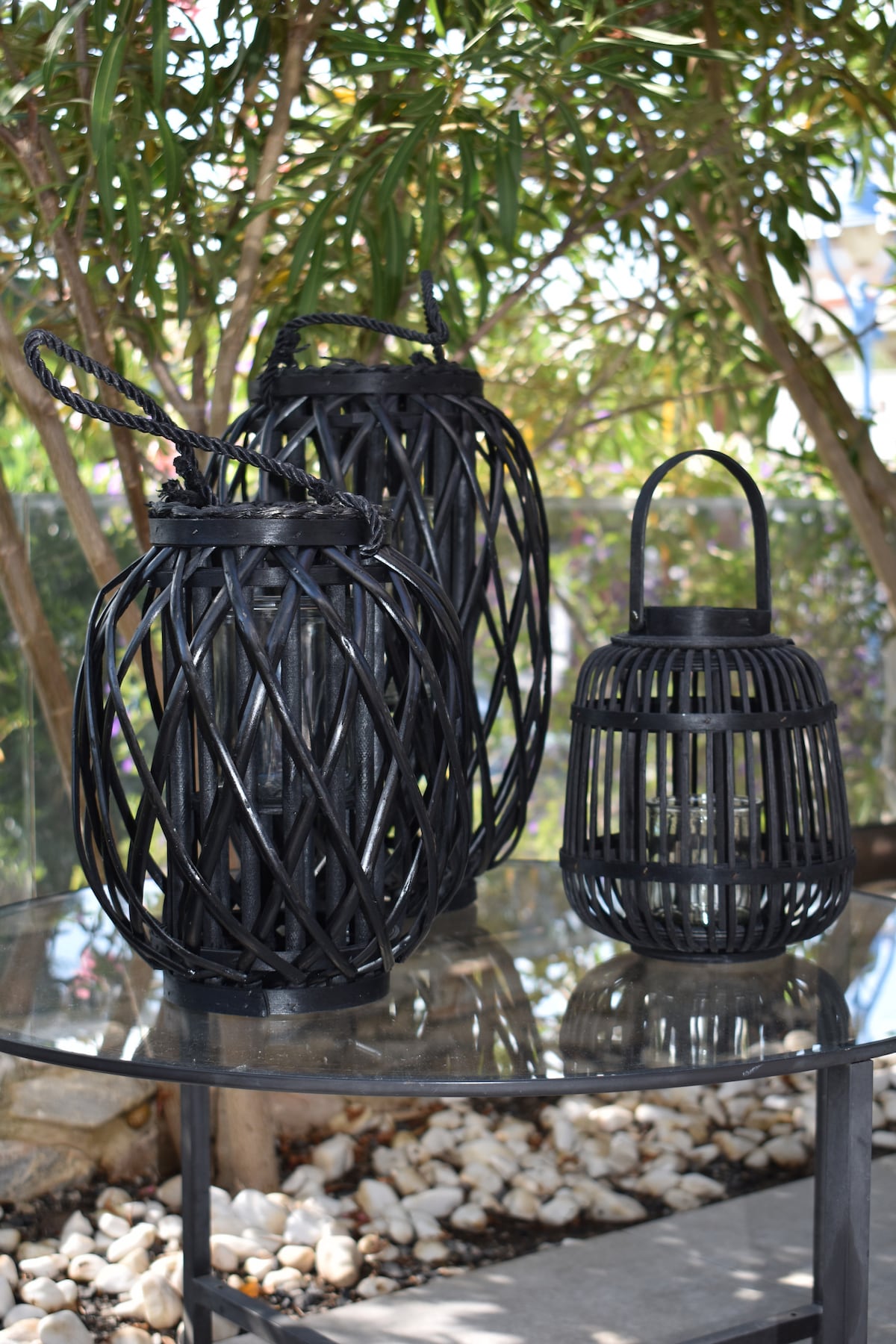 A glass tabletop is adorned with three decorative lanterns in various sizes, crafted from dark woven materials. The background features lush greenery, while smooth pebbles surround the base of the table, enhancing the outdoor setting.