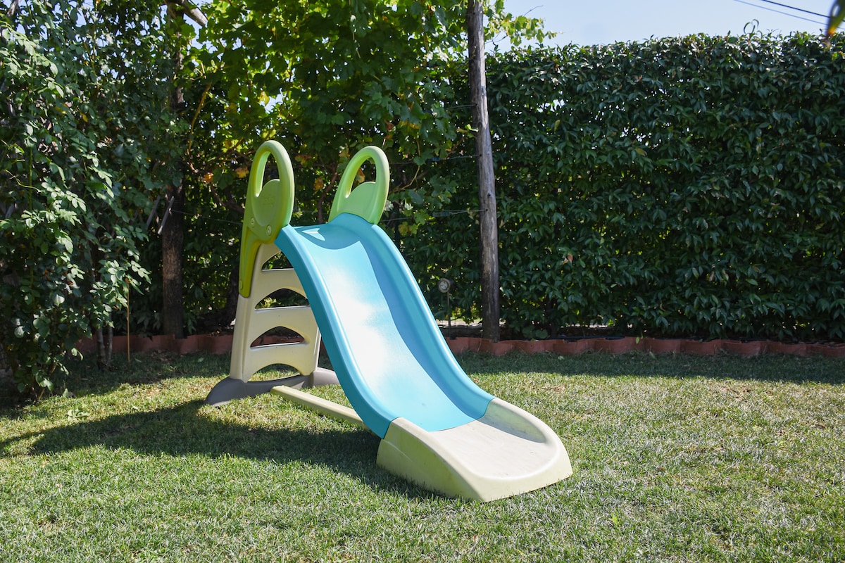 A colorful playground slide is placed on a grassy area, surrounded by lush green hedges. The slide features a gentle slope and sturdy handrails, designed for children's play.
