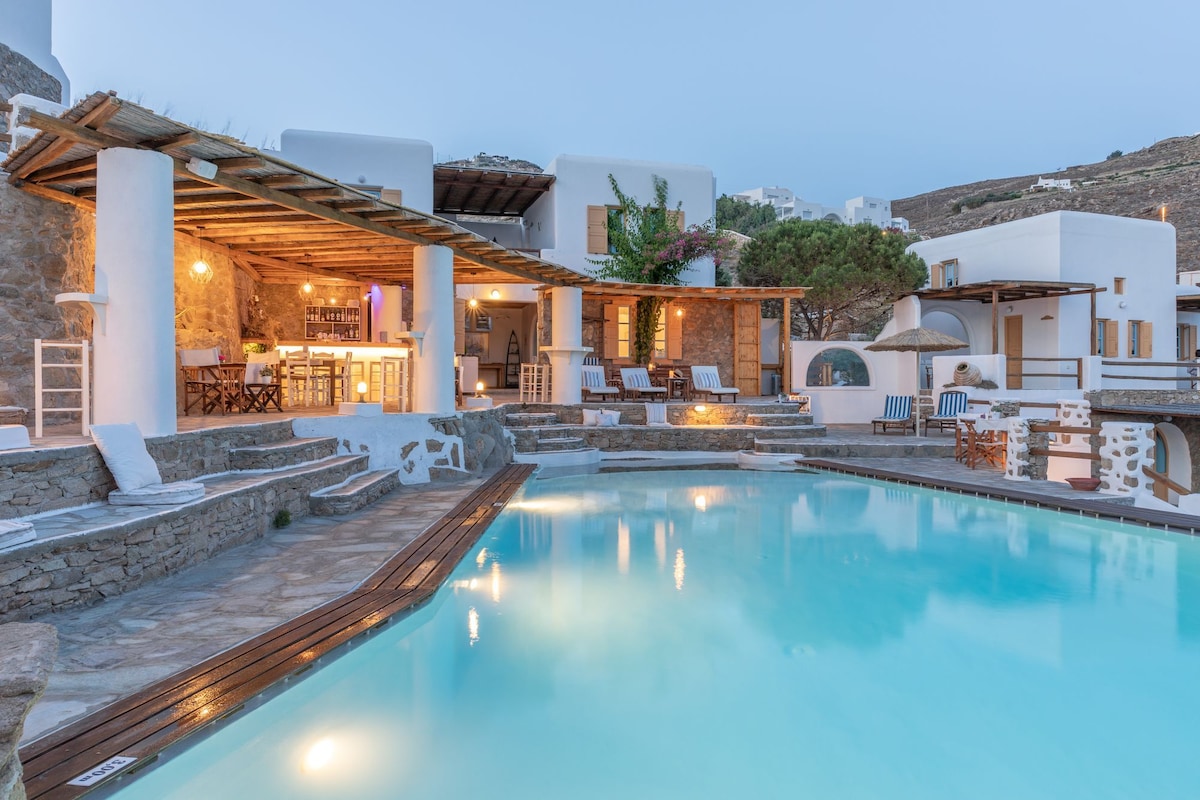 A shared swimming pool reflects soft blue tones under the evening sky, surrounded by a stone terrace. Light from an outdoor bar area enhances the warm ambiance of the traditional stone architecture. Comfortable lounge chairs are arranged nearby, inviting relaxation.