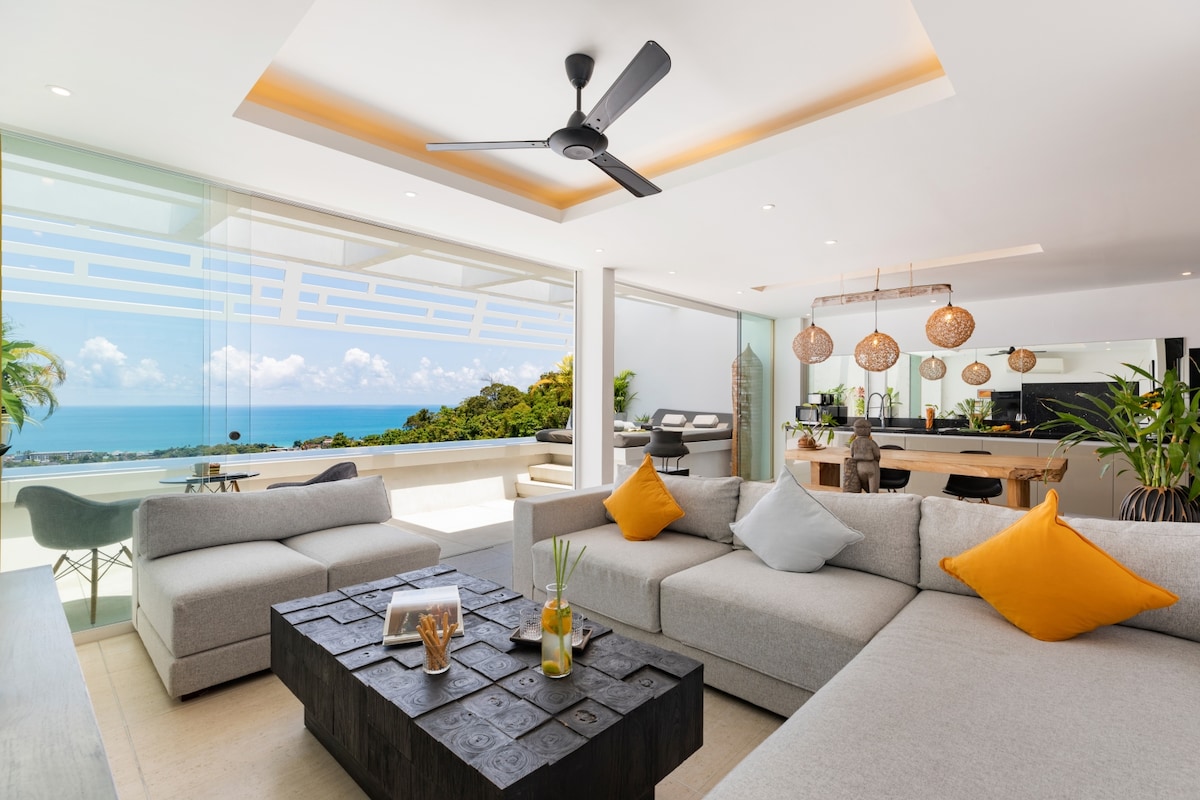 A modern living area is presented, featuring a spacious sectional sofa in light gray with vibrant orange and white cushions. A dark, textured coffee table sits at the center. Large glass panels frame a panoramic view of the sea, allowing ample natural light to illuminate the space.