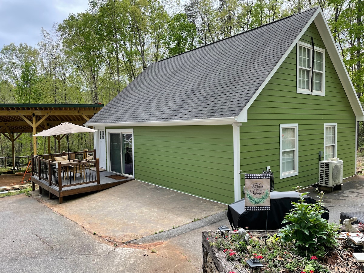 A green guest house is seen from the driveway, featuring a peaked roof and a front deck with outdoor seating. Trees surround the property, providing a natural backdrop. A sign indicating the property can be spotted near the flowerbed and a shaded area with a picnic table is visible.