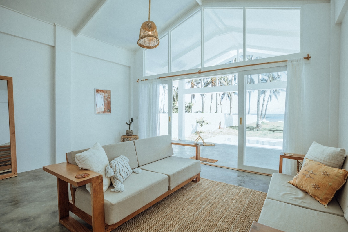A bright and airy living area features large glass sliding doors that open to the ocean view. Comfortable seating is arranged on a light-colored rug, with natural wood accents enhancing the space. Soft cushions are placed on the sofas, inviting relaxation in this open design.
