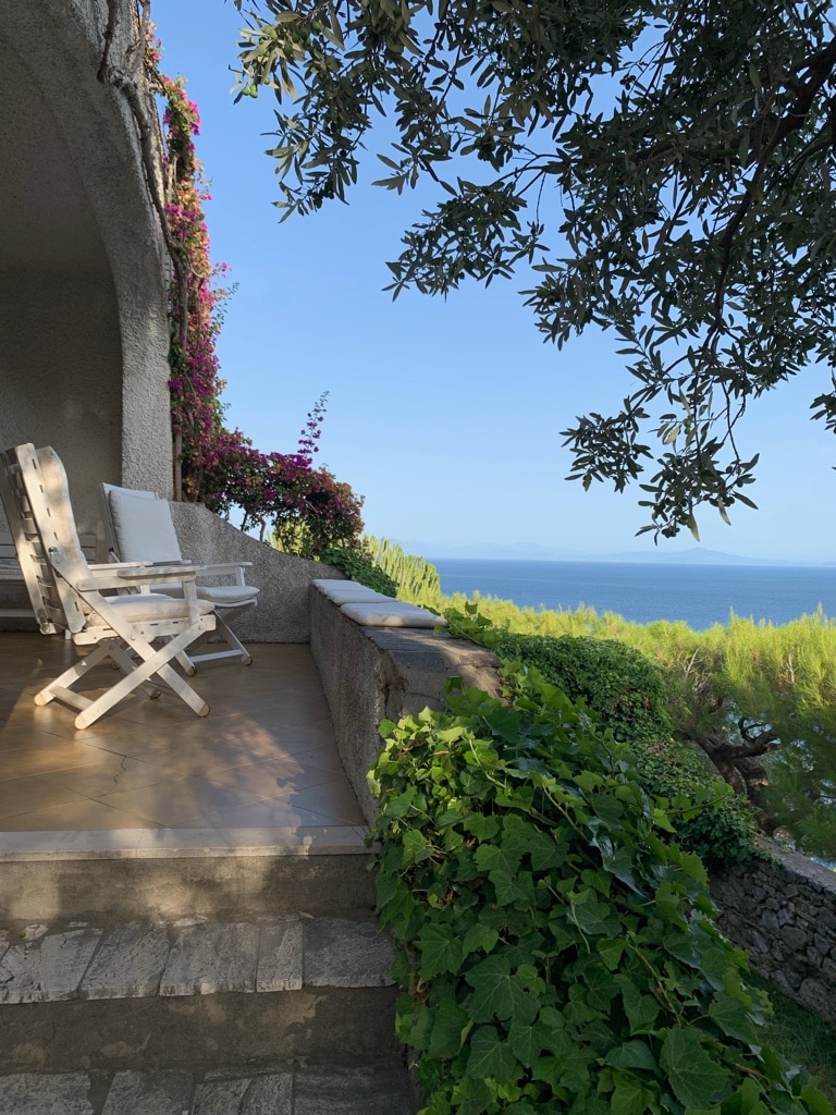 A charming terrace is framed by lush greenery and colorful flowers, offering a scenic view of the sea. Two white folding chairs are positioned on the wooden floor, inviting relaxation while enjoying the tranquil surroundings.