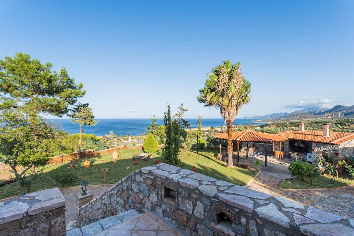 A view is presented overlooking the green landscape and coastline, with lush trees and tropical plants framing the scene. Terra-cotta rooftops of the villa are visible, accompanied by a stony pathway leading towards the garden and ocean beyond.