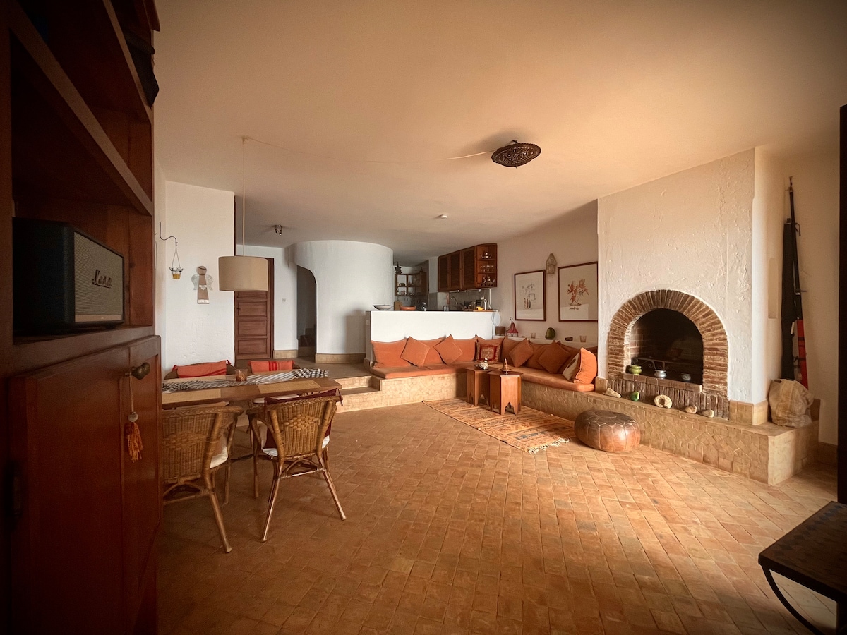 A spacious living room features a built-in fireplace framed by a rustic arch. Comfortable seating areas with orange cushions create an inviting space. Terracotta tiles cover the floor, and a wooden dining table is set nearby, offering a blend of comfort and warmth.