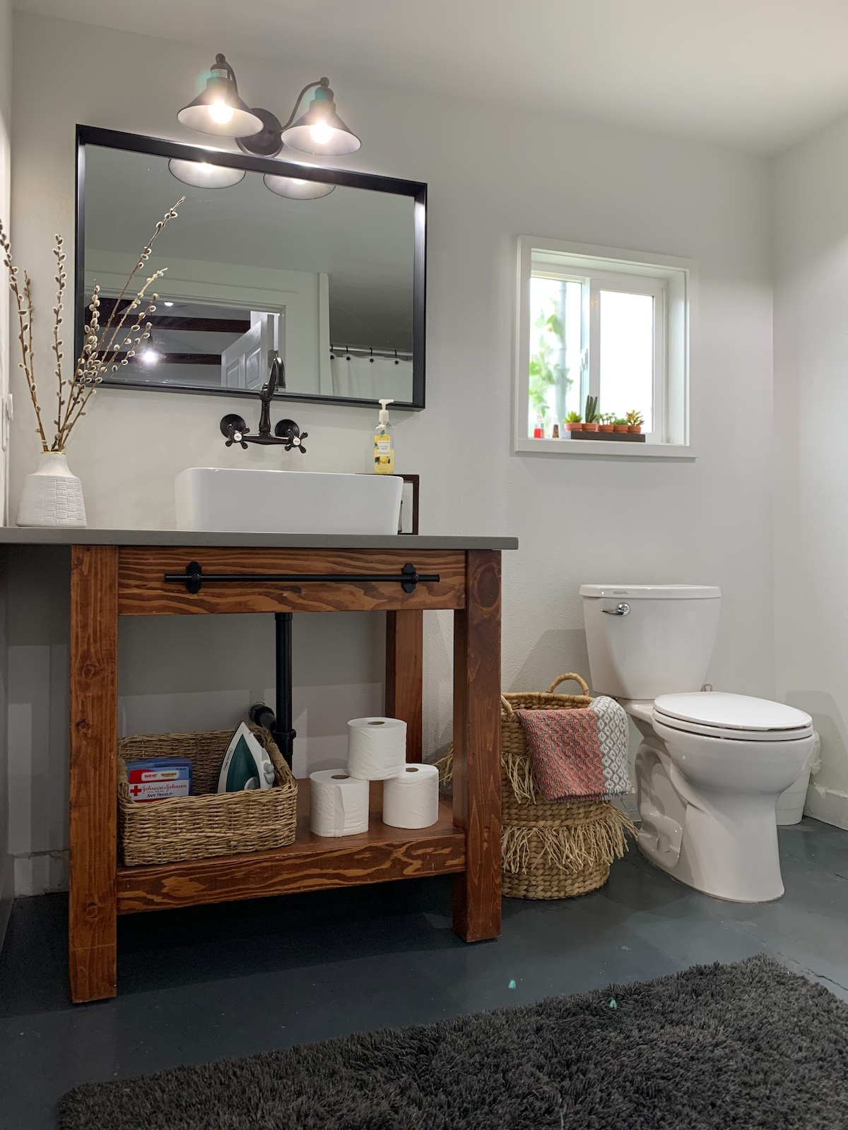 A bathroom is displayed with a wooden vanity featuring a sink and a large, framed mirror above. A small window allows natural light, while toiletries are organized on the countertop. A toilet is positioned adjacent to a basket, and bath towels are neatly arranged nearby.