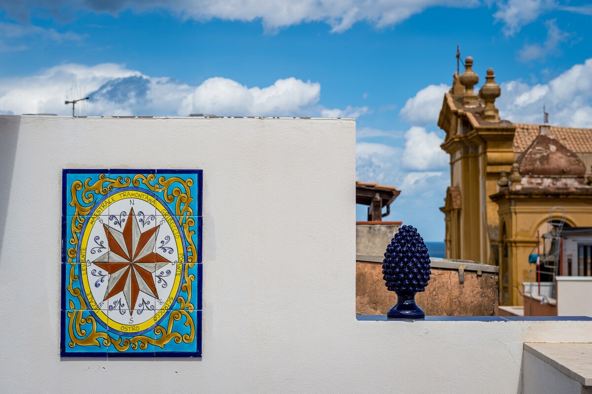 A vivid mosaic featuring a compass design is positioned against a white wall. A blue decorative object resembling a cluster of grapes rests nearby. The backdrop includes a building with a domed structure under a blue sky scattered with clouds.