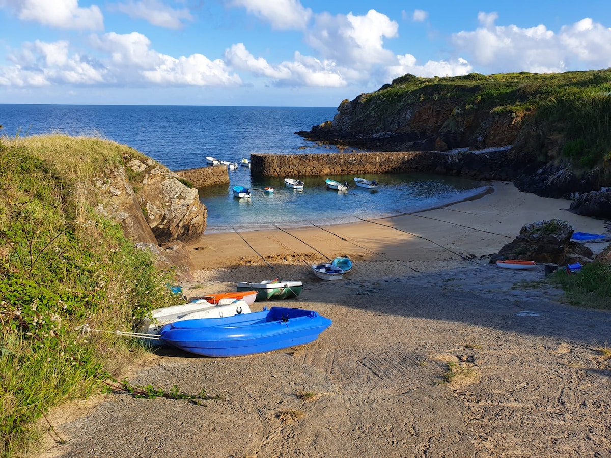 A calm, sandy cove is depicted, framed by rocky cliffs and gentle waves. Several small boats are moored at the shore, with vibrant colors enhancing the scene. The clear blue sky is dotted with clouds, reflecting a serene coastal environment.