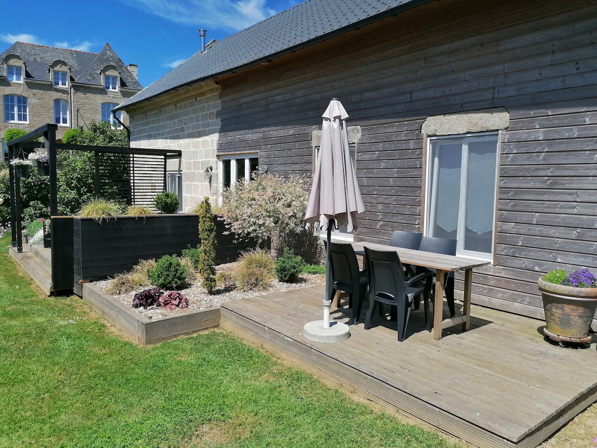 A wooden terrace features a dining table with four chairs and a large umbrella for shade. Surrounding areas are enhanced by raised garden beds with various plants, and the exterior of the building exhibits natural wood textures. Clear skies complement the outdoor space.