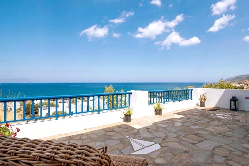 A spacious terrace is visible, featuring a natural stone floor and low blue railings that overlook the sea. Potted plants add greenery, while panoramic views of the water and sky can be seen in the background, creating a serene outdoor setting.