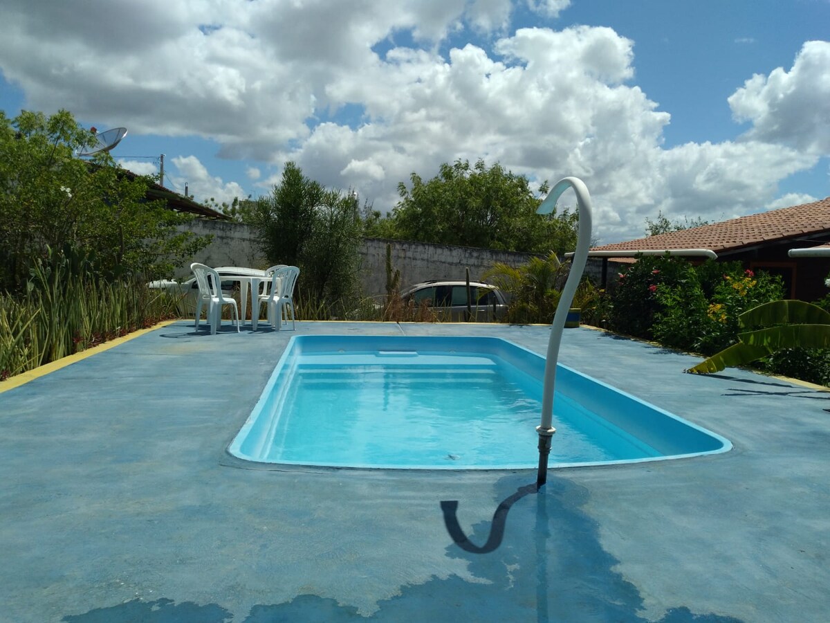 A small pool is situated on a concrete patio, surrounded by greenery and colorful shrubs. A set of white chairs and a table is positioned nearby. The sky above is partly cloudy, with several clouds visible.
