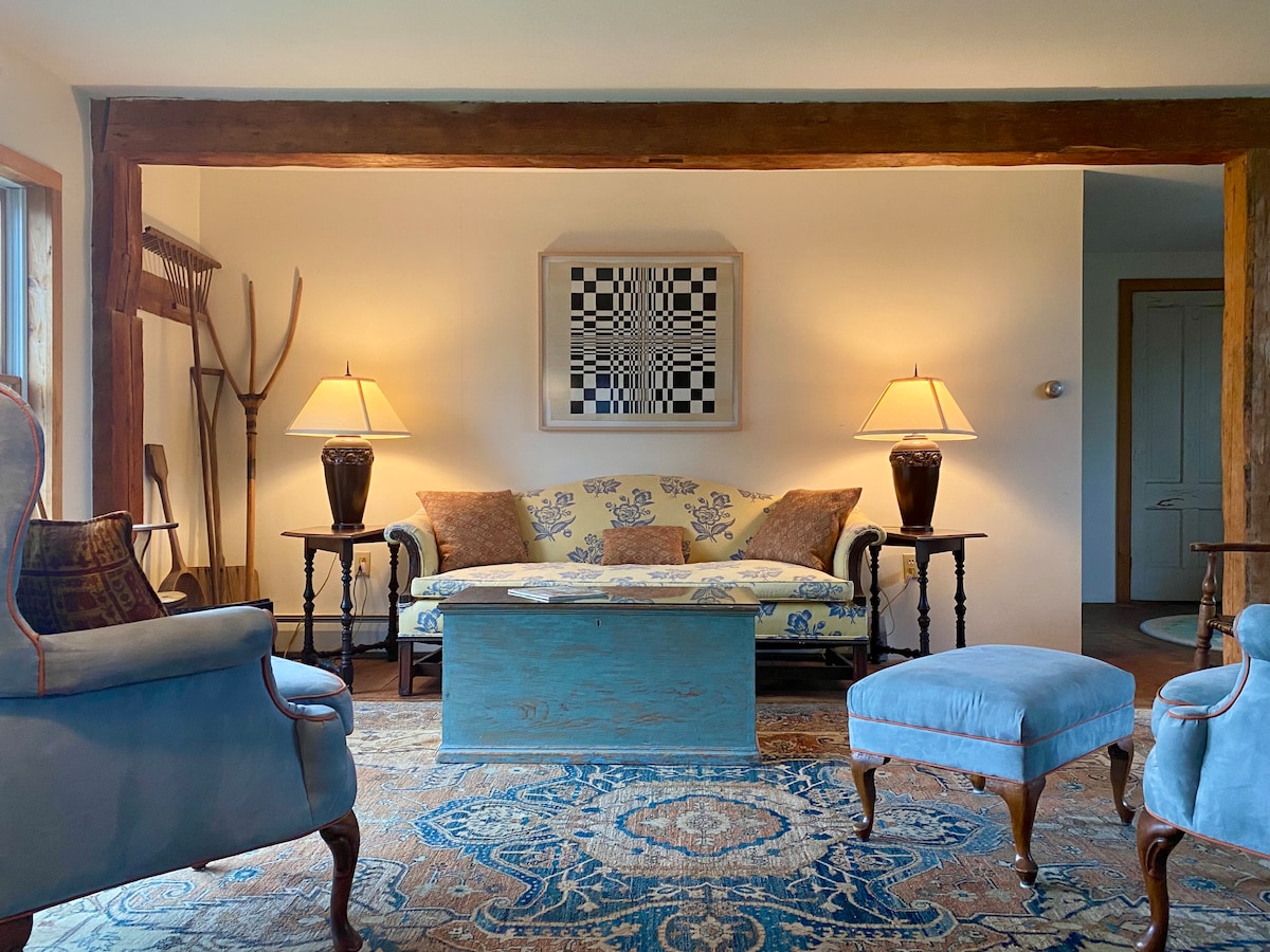 The living area features a floral-patterned sofa, complemented by two armchairs and a coordinating ottoman. A decorative blue coffee table is centered on a rich area rug, while two lamps illuminate the space from side tables. Exposed wooden beams add rustic charm.