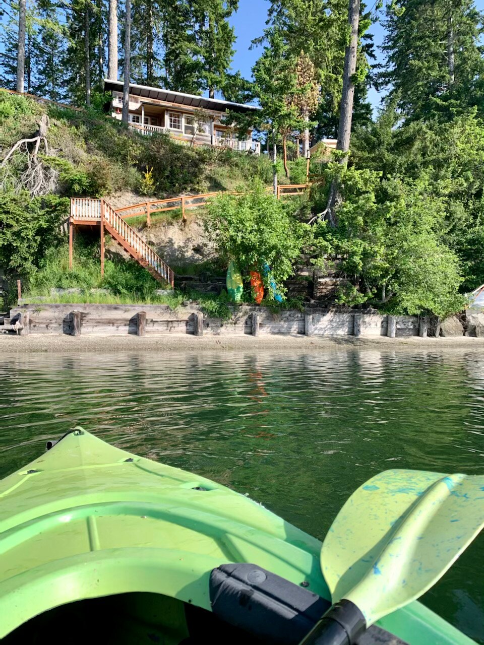A bright green kayak rests on calm waters, facing a wooden cabin perched on a hillside. A staircase with wooden railings leads up to the cabin, surrounded by lush greenery and trees. The shoreline features a smooth, sandy beach with visible rock formations.