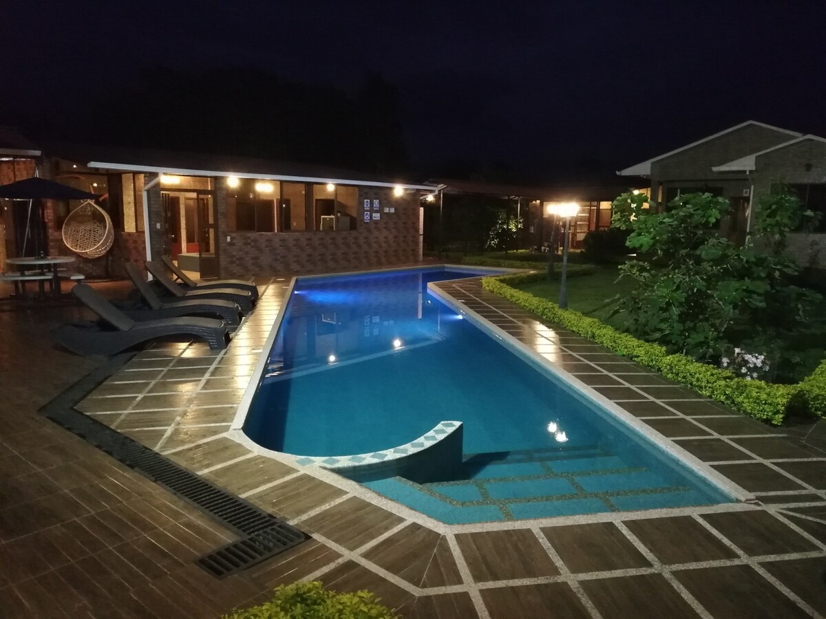 A well-lit swimming pool is featured, surrounded by dark wooden decking. Relaxation lounges are positioned alongside the pool, with lush greenery visible in the background. The area is enhanced by warm lighting, creating a welcoming space for guests to unwind.