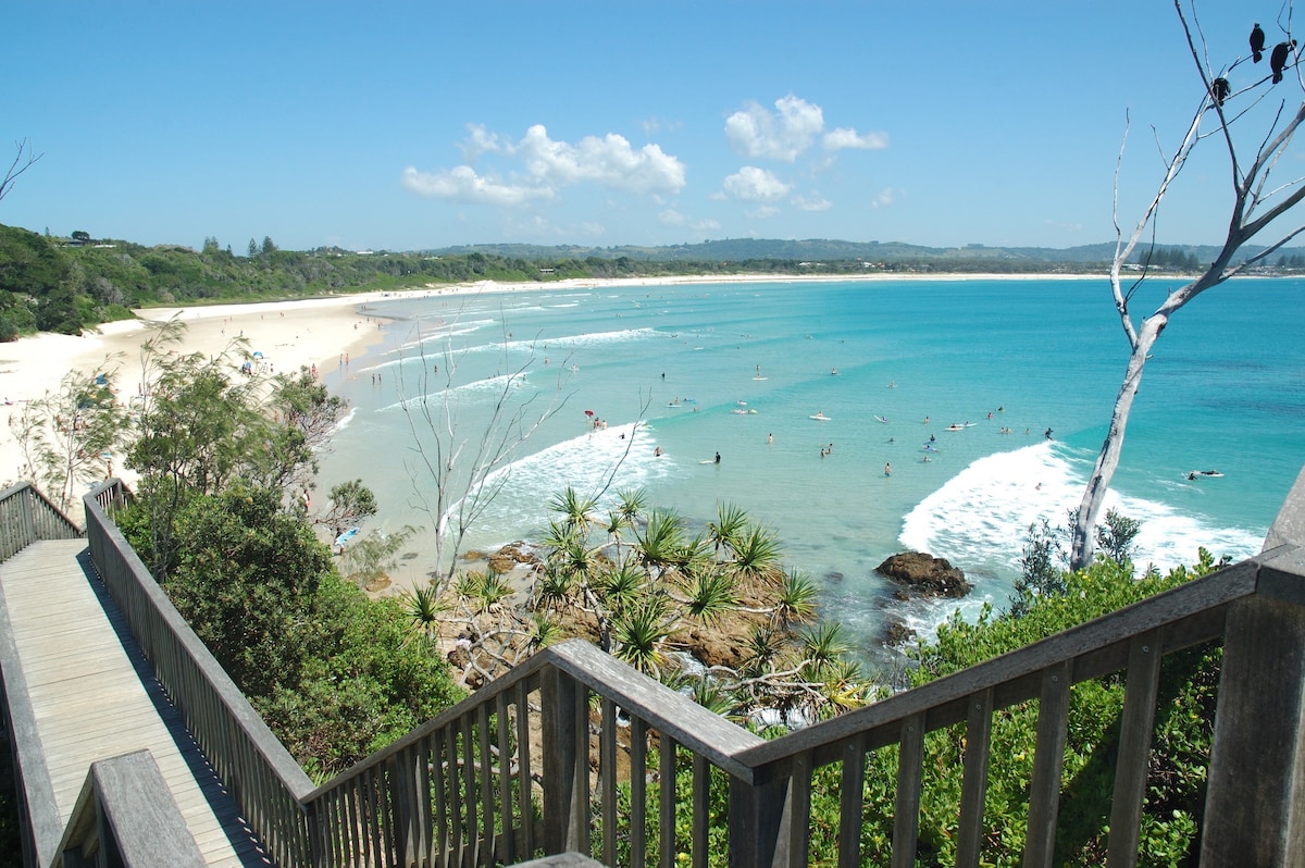 A panoramic view captures a serene beach scene, featuring golden sands meeting turquoise waters. Gentle waves lap at the shore, where visitors can be seen enjoying the water. Lush greenery frames the scene, with a wooden staircase leading down to the beach.