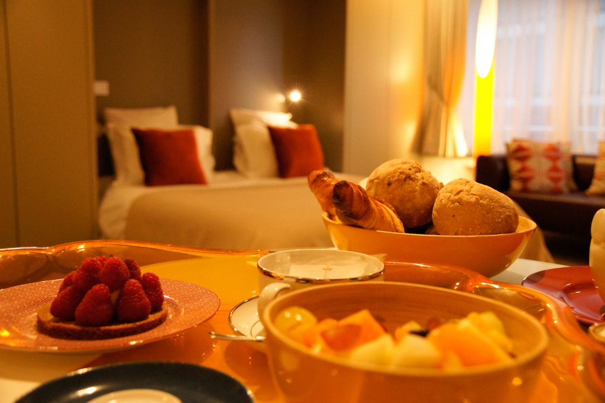 A close-up view of a breakfast spread features fresh raspberries, assorted pastries, and a colorful bowl of mixed fruit. In the background, a spacious bed with decorative pillows is positioned against a softly lit wall, offering a cozy atmosphere.