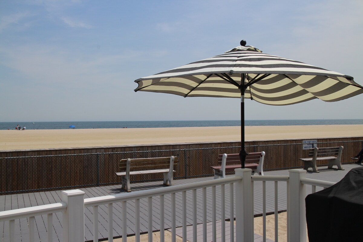 A striped umbrella provides shade over a wooden deck, which overlooks a wide expanse of pristine beach. Two benches face the water, where the ocean meets a clear blue sky. The calm sea is visible in the distance, with a few beachgoers enjoying the sand.