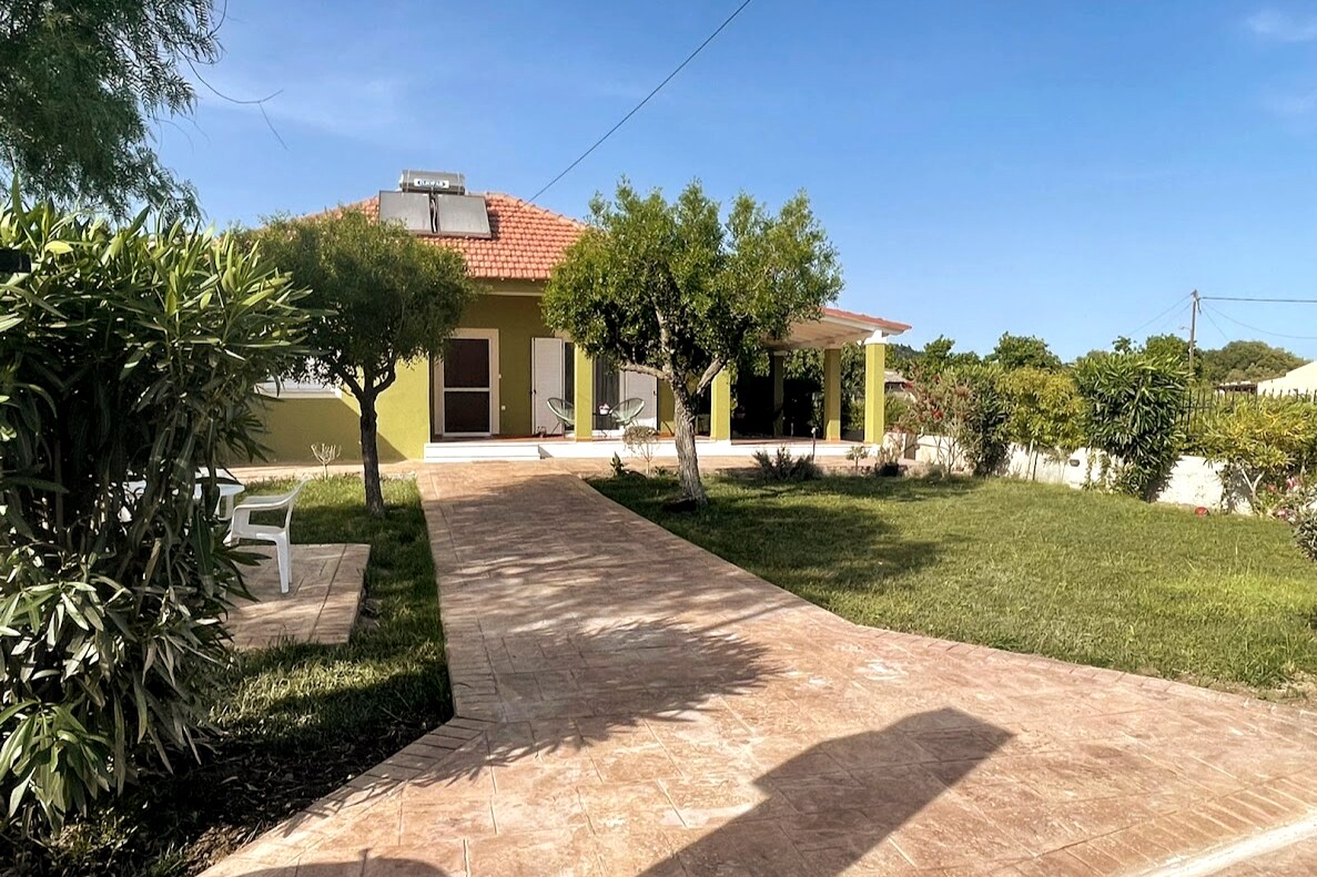 A country house is set against a clear blue sky, surrounded by well-kept gardens. A wide stone pathway leads to the entrance, flanked by lush greenery. The house features a red-tiled roof and large windows, inviting natural light into the living space.