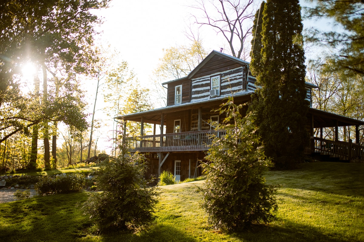 The historic log house is nestled among trees, featuring a wraparound porch that invites relaxation. Sunlight filters through the branches, casting a gentle glow on the surrounding green landscape. The rustic charm of the wooden exterior is complemented by well-maintained gardens.