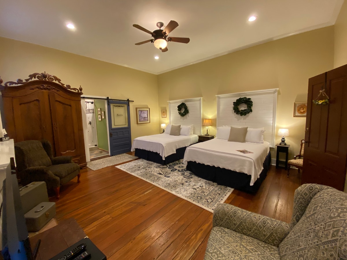 A spacious room features two beds with soft white linens set against light yellow walls. Antique furnishings, including a large wooden wardrobe and a comfortable armchair, complement the natural wood flooring. Two windows provide natural light, framed by decorative wreaths. A subtle area rug enhances the inviting ambiance.