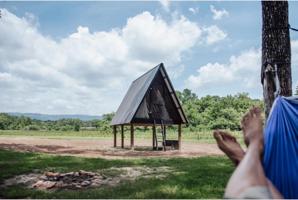 A rustic A-frame cabin with a sloped roof is positioned on wooden posts in an open field. A small set of stairs leads up to the entrance. A hammock is visible in the foreground, and the surrounding landscape features lush greenery and distant hills under partly cloudy skies.