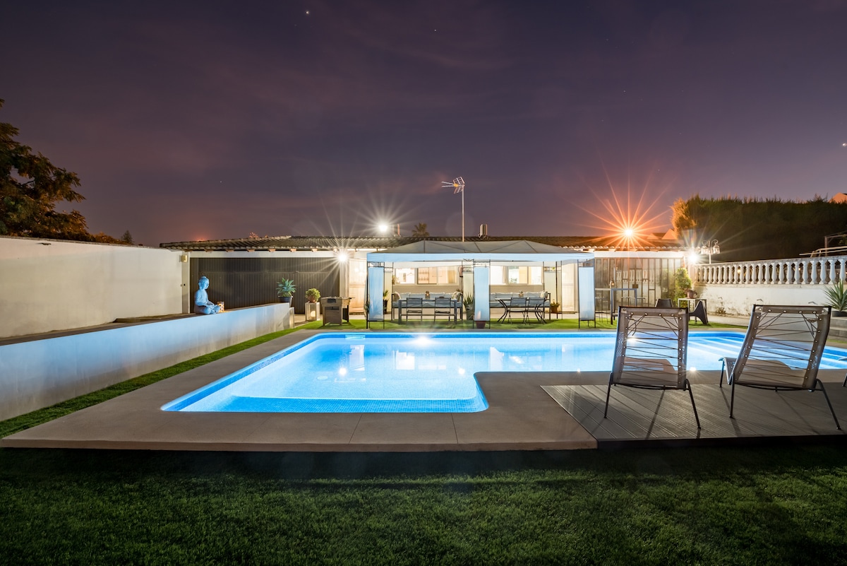 A spacious outdoor area features a well-lit swimming pool surrounded by a manicured lawn. Comfortable lounge chairs are positioned nearby, and a covered seating area with dining possibilities is visible in the background, enhancing the relaxation and enjoyment of the space.